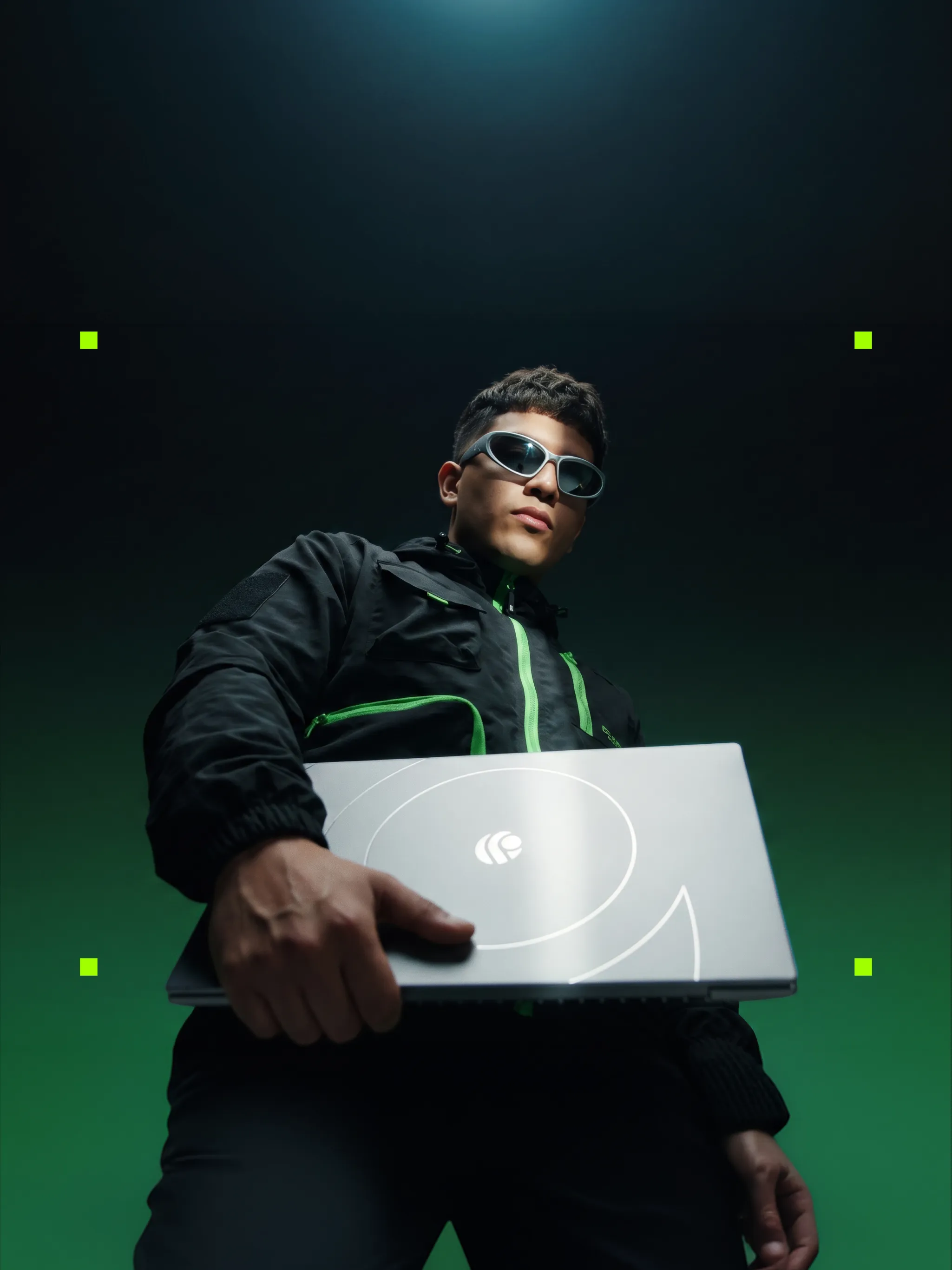 A young man in sunglasses and a dark jacket holding out a closed silver Primebook 3 laptop towards the camera